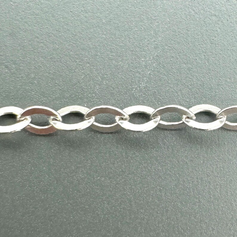 May include: Close-up of a silver-colored chain with oval links. The chain is made of a shiny, metallic material and is displayed against a gray background. The links are uniform in size and shape, creating a continuous pattern.
