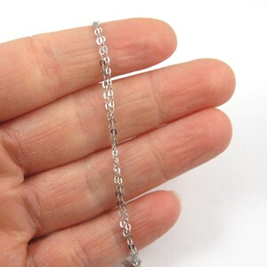 May include: A delicate silver chain necklace with a repeating oval link pattern. The chain is held in a person's hand against a white background. The chain is a fine, elegant piece of jewelry.