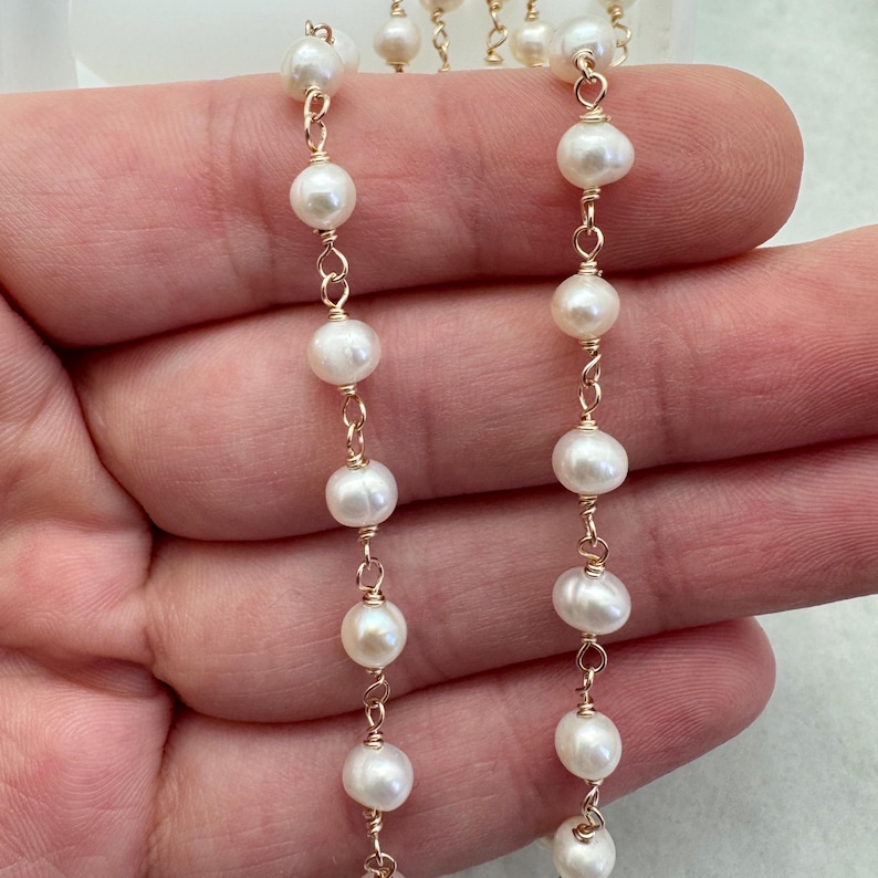Puede incluir: Primer plano de dos hilos de perlas blancas ensartadas en un delicado alambre dorado. Las perlas son redondas y est&aacute;n espaciadas uniformemente, creando un aspecto cl&aacute;sico y elegante. El fondo es de un color suave y neutro.