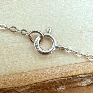 May include: Close-up of a silver necklace clasp and chain. The clasp is a circular design with a small lever. The chain is made of small, interconnected oval links. The background is a light brown wooden surface.
