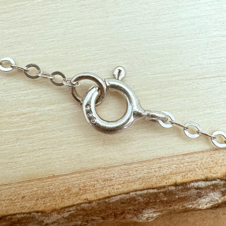 May include: Close-up of a silver necklace clasp and chain. The clasp is a circular design with a small lever. The chain is made of small, interconnected oval links. The background is a light brown wooden surface.