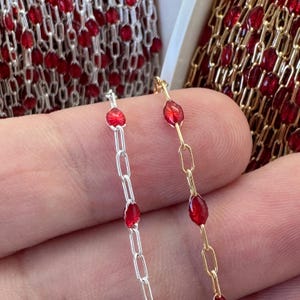 May include: Close-up of two chains with red gemstone beads. One chain is silver-toned, the other is gold-toned. The chains are linked with small, rectangular links. The red beads are oval-shaped and are evenly spaced along the chains. The background shows more chains.