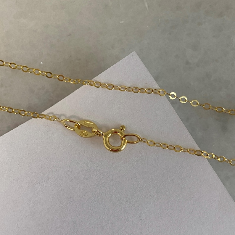 May include: A gold-colored metal chain necklace featuring a circular spring ring clasp and an oval-shaped jump ring connector. The chain is composed of small, uniform, interconnected oval links that create a delicate and continuous strand design.