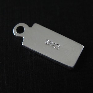 Puede incluir: Encanto de plata con forma rectangular y un bucle en la parte superior. El encanto está marcado con el número "925", lo que indica que está hecho de plata de ley.