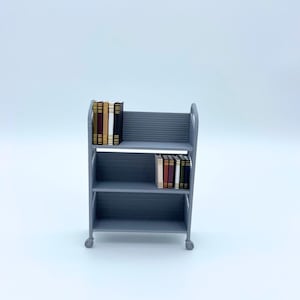 Peut inclure: Une étagère grise à trois niveaux sur roues, contenant plusieurs livres miniatures. Les étagères sont horizontales et les livres ont des dos de couleurs variées. L'étagère est un modèle miniature, probablement pour une maison de poupée ou une exposition.