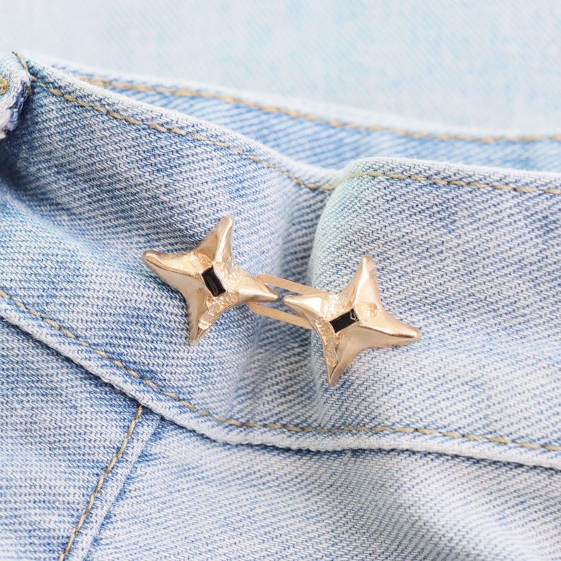 Puede incluir: Un par de pendientes dorados en forma de estrella con detalles negros. Los pendientes se colocan sobre un fondo de mezclilla azul claro, resaltando el dise&ntilde;o y el contraste de color de la joya. Un accesorio de moda.