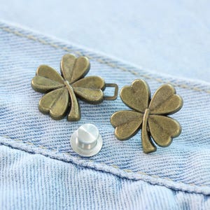 Może przedstawiać: Dwa guziki w kształcie koniczyny w kolorze brązu i srebrny guzik są wyświetlane na jasnoniebieskim tle z dżinsu. Guziki w kształcie koniczyny mają szczeg&oacute;łowy, teksturowany wz&oacute;r, a guzik jest okrągły.