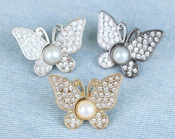 May include: Three butterfly brooches. Two are silver-toned, one with clear crystals and the other with a pearl center. The third brooch is gold-toned, also with clear crystals and a pearl center. All three are set against a light blue background.