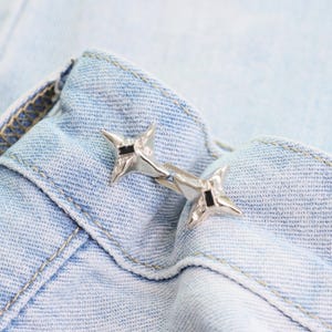 Puede incluir: Un broche de metal plateado con un dise&ntilde;o de estrella y detalles en negro. El broche est&aacute; sujeto a una tela vaquera azul claro, mostrando la textura del material y los detalles intrincados.
