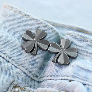 Może przedstawiać: Dwa srebrne guziki w kształcie koniczyny są prezentowane na jasnoniebieskim dżinsie. Guziki mają metaliczny połysk i są umieszczone w pobliżu paska dżins&oacute;w. Koniczyna to klasyczny symbol szczęścia.