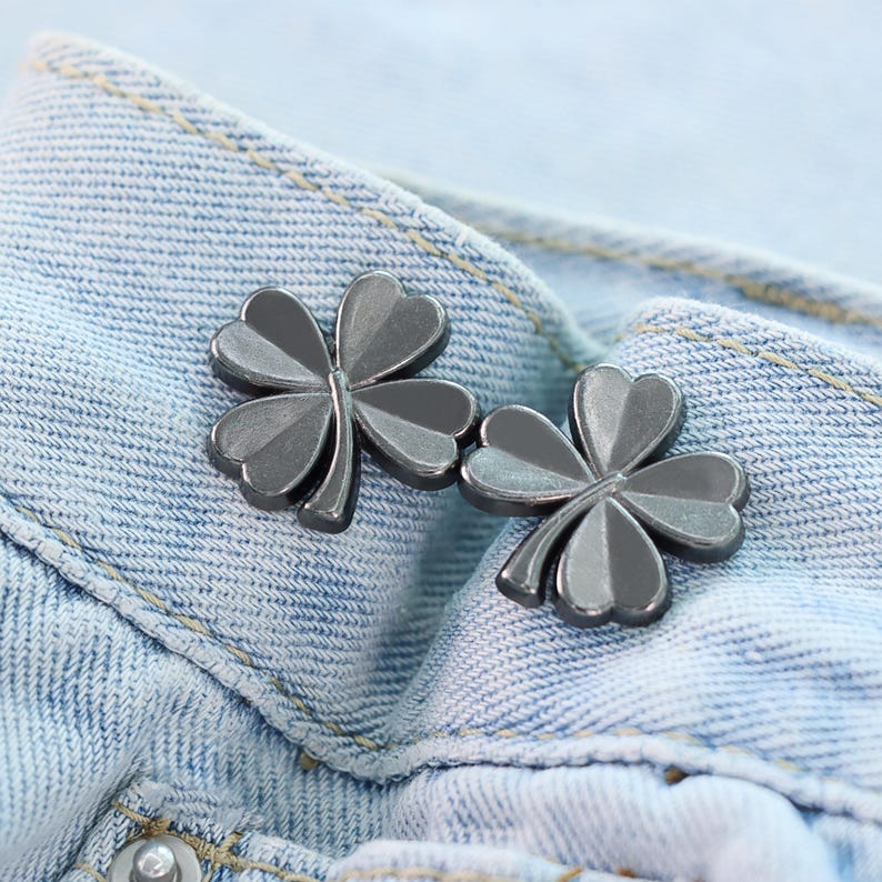 Może przedstawiać: Dwa srebrne guziki w kształcie koniczyny są prezentowane na jasnoniebieskim dżinsie. Guziki mają metaliczny połysk i są umieszczone w pobliżu paska dżins&oacute;w. Koniczyna to klasyczny symbol szczęścia.