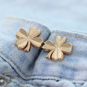 Może przedstawiać: Dwa złote guziki w kształcie koniczyny są prezentowane na jasnoniebieskim tle z dżinsu. Guziki mają wz&oacute;r czterolistnej koniczyny, z każdym liściem szczeg&oacute;łowym i lekko zakrzywionym.