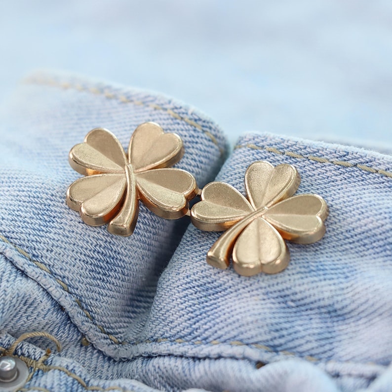 Może przedstawiać: Dwa złote guziki w kształcie koniczyny są prezentowane na jasnoniebieskim tle z dżinsu. Guziki mają wz&oacute;r czterolistnej koniczyny, z każdym liściem szczeg&oacute;łowym i lekko zakrzywionym.