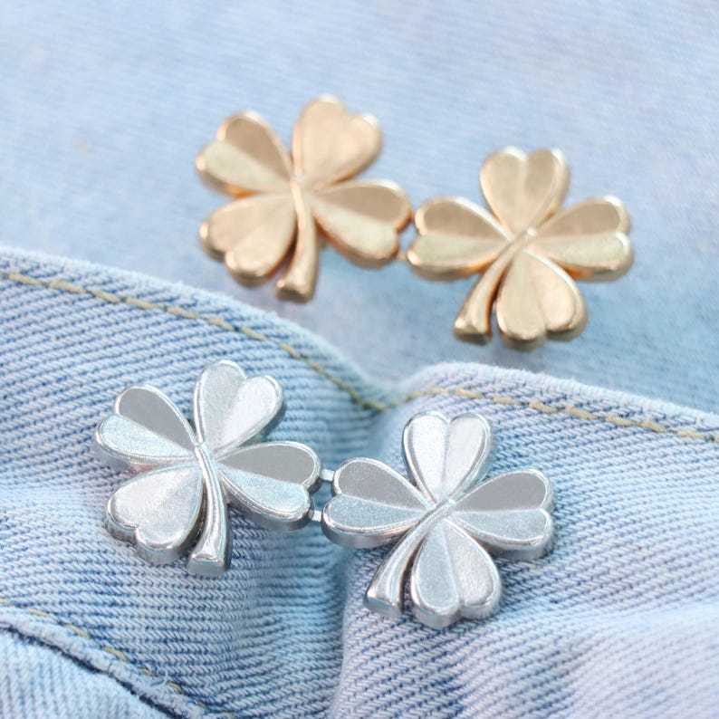 Może przedstawiać: Dwa zestawy guzik&oacute;w w kształcie koniczyny w odcieniach złota i srebra są prezentowane na niebieskim tle z dżinsu. Guziki są wykonane z metalu i mają tr&oacute;jwymiarowy design. Liście koniczyny są szczeg&oacute;łowe z zakrzywionymi krawędziami.