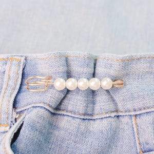 May include: A decorative pearl pin, featuring a row of five white pearls, is attached to the waistband of a light blue denim garment. The pin has a silver-colored clasp and a straight pin design, adding a touch of elegance to the casual fabric.