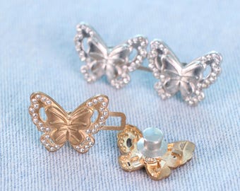 May include: Close-up of three butterfly-shaped metal buttons. Two are silver-colored, and one is gold-colored. Each butterfly has a detailed wing design and is embellished with small, clear rhinestones. The buttons are on a light blue fabric.