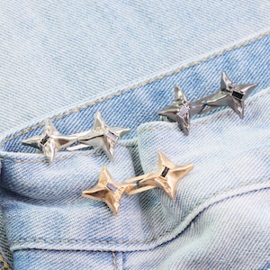 Puede incluir: Tres pares de anillos en forma de estrella en plata, oro y metal de ca&ntilde;&oacute;n, cada uno con un acento cuadrado negro. Los anillos se muestran sobre una tela vaquera azul claro, mostrando sus variaciones de dise&ntilde;o y color.