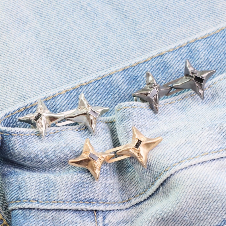 Puede incluir: Tres pares de anillos en forma de estrella en plata, oro y metal de ca&ntilde;&oacute;n, cada uno con un acento cuadrado negro. Los anillos se muestran sobre una tela vaquera azul claro, mostrando sus variaciones de dise&ntilde;o y color.