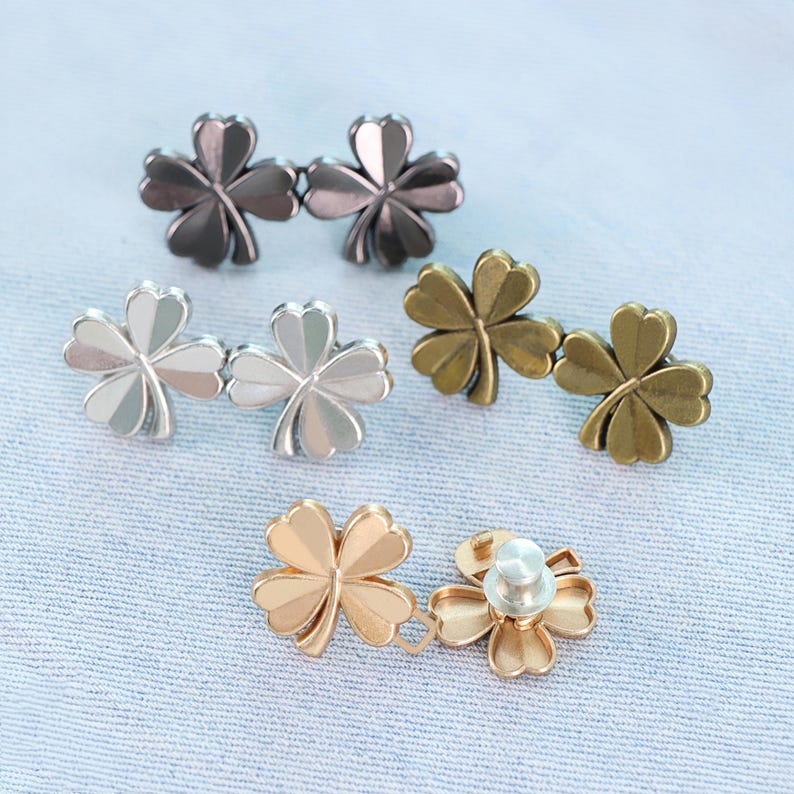 Może przedstawiać: Kolekcja metalowych guzik&oacute;w w kształcie koniczyny w r&oacute;żnych wykończeniach: grafitowym, srebrnym, antycznym mosiądzu i złotym. Guziki ułożone są na jasnoniebieskim tle z tkaniny, prezentując ich r&oacute;żne kolory i wzory.