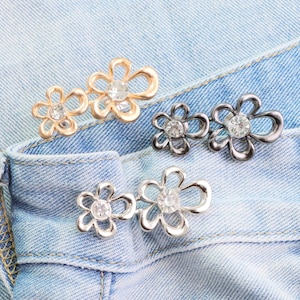 Puede incluir: Primer plano de varios botones decorativos con forma de flor y centros de pedrería. Los botones son en tonos dorados y plateados, y están sujetos a una tela vaquera azul claro. Estos botones se pueden utilizar para proyectos de costura o manualidades.