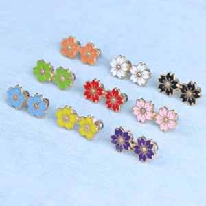 Puede incluir: Una colección de pendientes de botón con forma de flor en varios colores. Los pendientes están disponibles en azul, verde, naranja, blanco, rojo, negro, rosa, amarillo y morado. Cada pendiente tiene un respaldo de metal dorado.