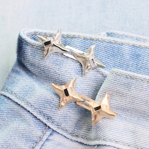 Puede incluir: Dos horquillas met&aacute;licas en forma de estrella, una plateada y otra dorada, descansan sobre el borde doblado de una prenda vaquera azul claro. Cada horquilla presenta un acento cuadrado negro en el centro de la estrella.