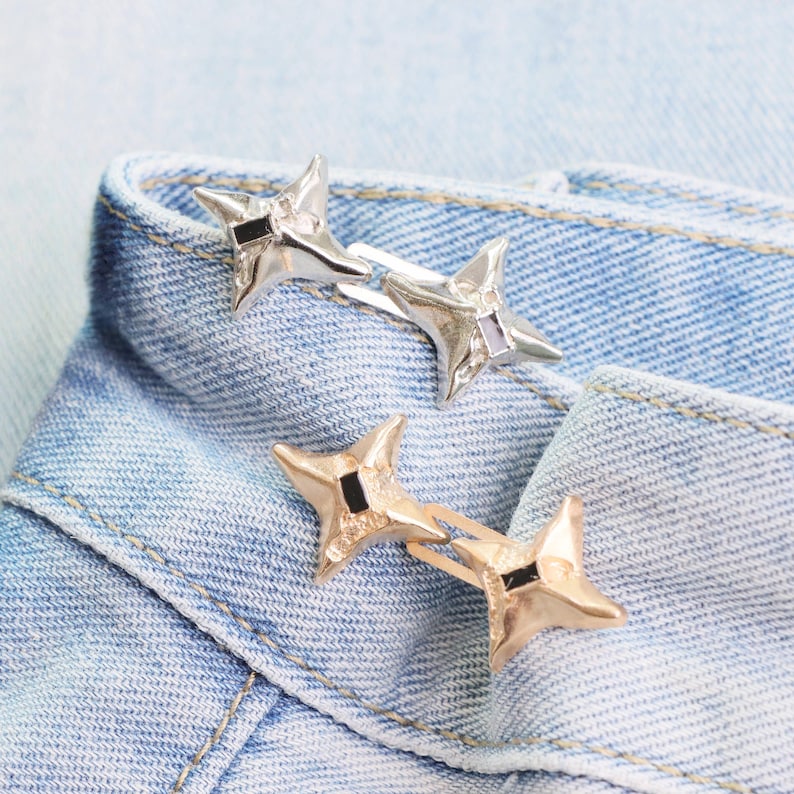Puede incluir: Dos horquillas met&aacute;licas en forma de estrella, una plateada y otra dorada, descansan sobre el borde doblado de una prenda vaquera azul claro. Cada horquilla presenta un acento cuadrado negro en el centro de la estrella.
