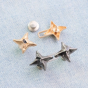 Puede incluir: Colecci&oacute;n de tachuelas met&aacute;licas en forma de estrella en colores oro y metal de ca&ntilde;&oacute;n. Una tachuela tiene un respaldo de alfiler, otras tienen respaldos de rosca. Las tachuelas se muestran sobre una superficie de mezclilla azul claro.