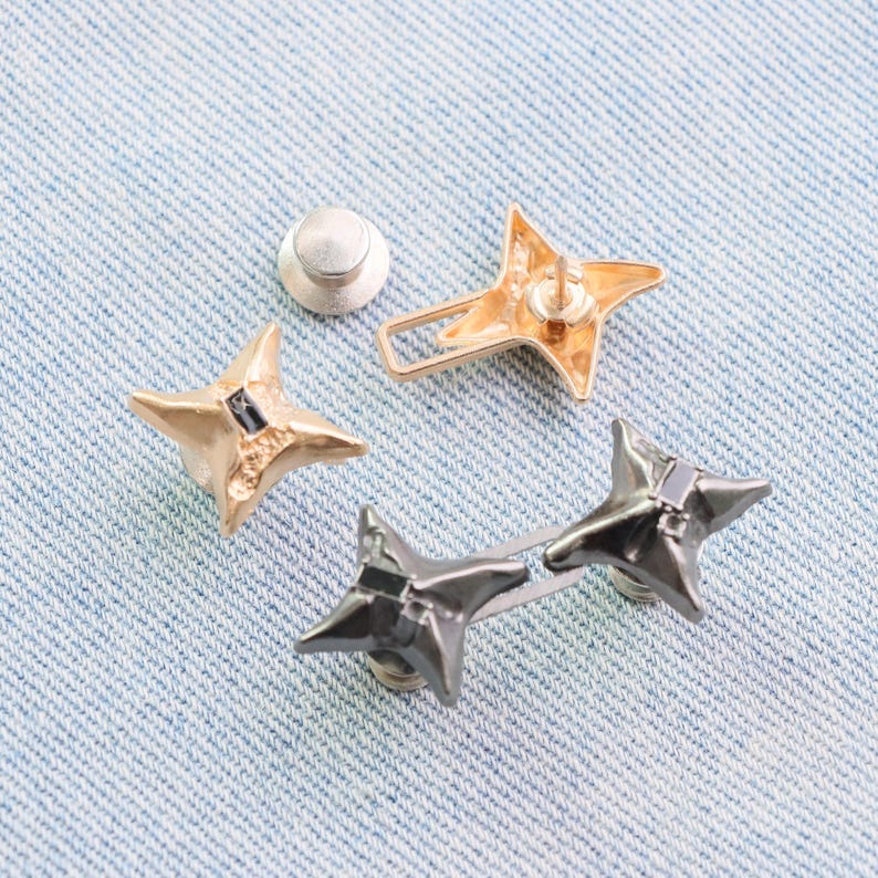 Puede incluir: Colecci&oacute;n de tachuelas met&aacute;licas en forma de estrella en colores oro y metal de ca&ntilde;&oacute;n. Una tachuela tiene un respaldo de alfiler, otras tienen respaldos de rosca. Las tachuelas se muestran sobre una superficie de mezclilla azul claro.