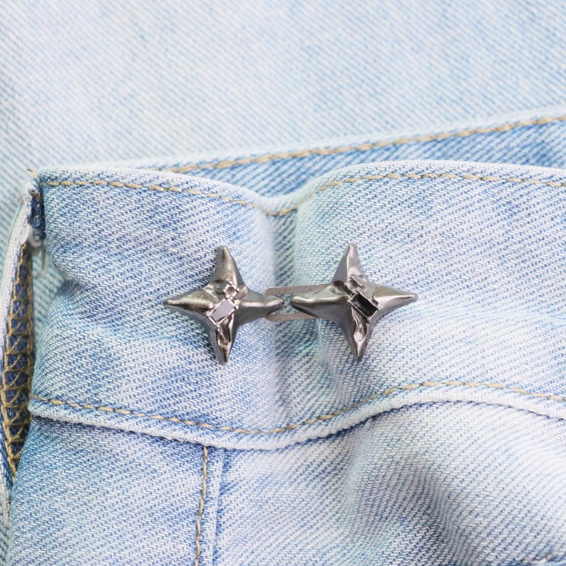 Puede incluir: Primer plano de dos remaches met&aacute;licos en forma de estrella sobre tela vaquera azul claro. Los remaches son de color met&aacute;lico oscuro y tienen un dise&ntilde;o de estrella de cuatro puntas. El denim tiene costuras visibles.