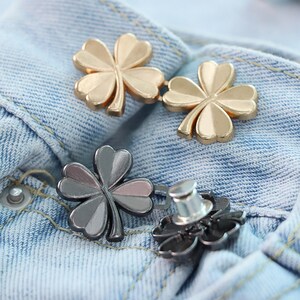 Może przedstawiać: Zbliżenie metalowych guzik&oacute;w w kształcie czterolistnej koniczyny w odcieniach złota i srebra. Guziki są przymocowane do jasnoniebieskiego dżinsu. Guziki mają zapięcie na szpilkę.