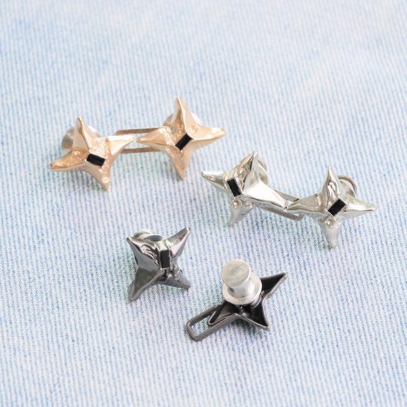 Puede incluir: Cuatro tachuelas met&aacute;licas en forma de estrella en oro, plata y negro. Cada estrella tiene cuatro puntas y un peque&ntilde;o cuadrado negro en el centro. Dos tachuelas est&aacute;n conectadas por una barra. Las tachuelas est&aacute;n sobre un fondo de mezclilla azul claro.