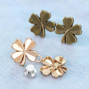 Może przedstawiać: Metalowe elementy biżuterii w kształcie czterolistnej koniczyny w odcieniach złota i brązu. Jedna złota koniczyna ma białe emaliowane akcenty. Elementy obejmują szpilkę i oddzielne zapięcie. Koniczyny są prezentowane na jasnoniebieskiej tkaninie.