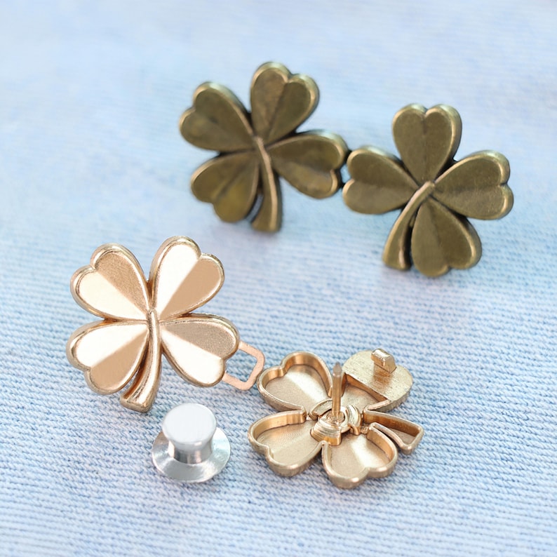 Może przedstawiać: Metalowe elementy biżuterii w kształcie czterolistnej koniczyny w odcieniach złota i brązu. Jedna złota koniczyna ma białe emaliowane akcenty. Elementy obejmują szpilkę i oddzielne zapięcie. Koniczyny są prezentowane na jasnoniebieskiej tkaninie.