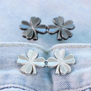 Może przedstawiać: Dwa zestawy srebrnych zapięć w kształcie koniczyny na jasnoniebieskim tle z dżinsu. Zapięcia są wykonane z metalu i mają czterolistny wz&oacute;r koniczyny. Tkanina dżinsowa ma widoczne szwy.