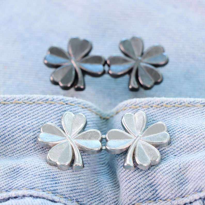 Może przedstawiać: Dwa zestawy srebrnych zapięć w kształcie koniczyny na jasnoniebieskim tle z dżinsu. Zapięcia są wykonane z metalu i mają czterolistny wz&oacute;r koniczyny. Tkanina dżinsowa ma widoczne szwy.