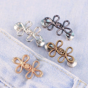 Könnte beinhalten: Vier Sets dekorativer blütenförmiger Knöpfe in Silber, Gunmetal, Bronze und Gold. Jeder Knopf hat ein Blumendesign mit einem Strass-Mittelteil und einer Metallrückseite. Die Knöpfe werden auf einem hellblauen Denim-Stoff präsentiert.