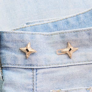 Puede incluir: Primer plano de dos botones de jean dorados en forma de estrella sobre mezclilla azul claro. Cada bot&oacute;n tiene un peque&ntilde;o cuadrado negro en el centro. Los botones est&aacute;n sujetos a la cintura de los jeans.