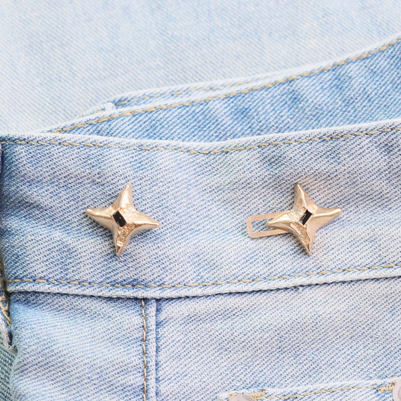 Puede incluir: Primer plano de dos botones de jean dorados en forma de estrella sobre mezclilla azul claro. Cada bot&oacute;n tiene un peque&ntilde;o cuadrado negro en el centro. Los botones est&aacute;n sujetos a la cintura de los jeans.