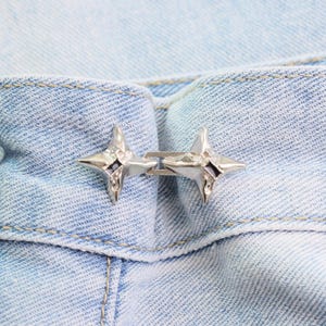Puede incluir: Primer plano de un cierre de metal plateado con dise&ntilde;o de estrella. El cierre est&aacute; sujeto a una tela vaquera azul claro. Las estrellas tienen un centro oscuro y est&aacute;n conectadas por una peque&ntilde;a barra de metal.