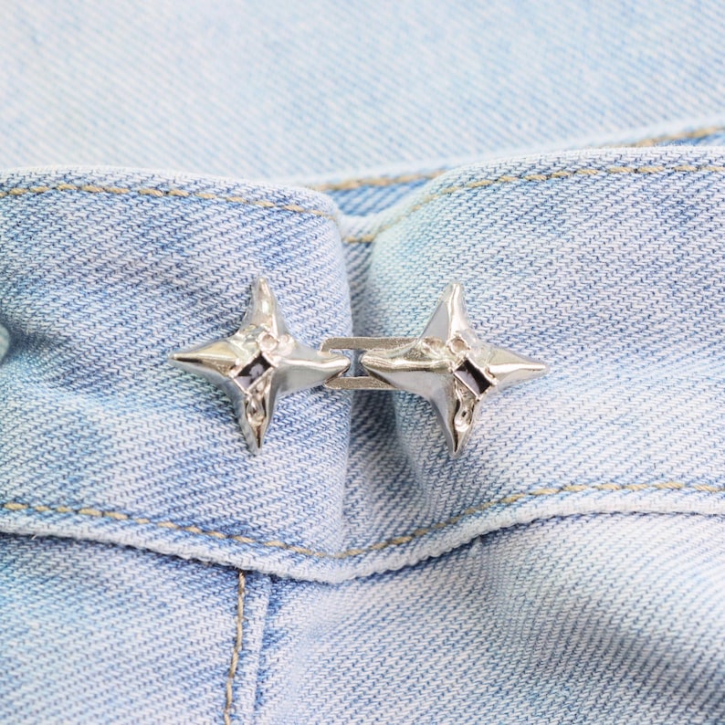 Puede incluir: Primer plano de un cierre de metal plateado con dise&ntilde;o de estrella. El cierre est&aacute; sujeto a una tela vaquera azul claro. Las estrellas tienen un centro oscuro y est&aacute;n conectadas por una peque&ntilde;a barra de metal.