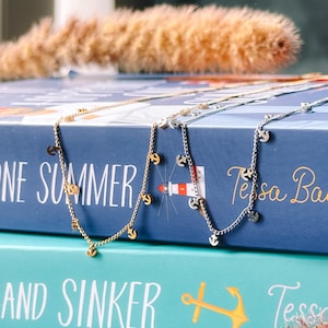 May include: Two delicate gold and silver necklaces with anchor charms, displayed on a stack of books with the titles "One Summer" and "And Sinker" visible.