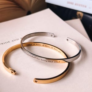 Puede incluir: Dos pulseras de brazalete de metal, una dorada y otra plateada, se muestran en una página de libro. La pulsera dorada tiene la inscripción "I have bewitched me. body and soul". La pulsera plateada tiene la inscripción "and 9 love, love, love you."
