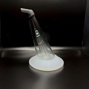 Puede incluir: Una pipa de agua de vidrio transparente con forma cónica, con boquilla curva y base circular blanca. El interior tiene un diseño texturizado y se aprecia un pequeño acento verde. La pipa está inclinada, mostrando su diseño sobre un fondo oscuro.