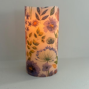 May include: A cylindrical candle with a floral design. The candle features a watercolor pattern of purple, orange, and green flowers and leaves. The candle is lit, emitting a warm, orange glow. The candle is sitting on a white surface.