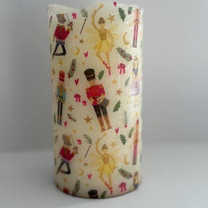May include: A cream-colored pillar candle featuring a repeating pattern of Nutcracker soldiers, fairies, stars, and bows. The candle has a slightly wavy top edge and is designed for decorative use.