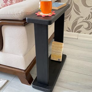 Può includere: Un tavolino nero in legno con piano rettangolare e ripiano inferiore. Una tazza arancione è appoggiata su un sottobicchiere decorativo, accanto a un libro. Il tavolo è accanto a un divano beige con un cuscino floreale.