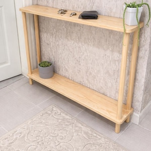 Op de afbeelding: Een lichtgekleurde houten console met twee planken. De bovenste plank bevat sleutels, een portemonnee en een kleine plant in een pot. Een tweede plant in een pot staat op de onderste plank. Een patroonkleed staat op de voorgrond.