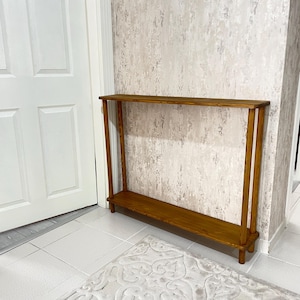 Op de afbeelding: Een houten consoletafel met een natuurlijke houten afwerking. De tafel heeft &eacute;&eacute;n plank en wordt ondersteund door vier poten.