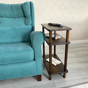 Könnte beinhalten: Ein dunkelbrauner Beistelltisch mit drei Ebenen, abgerundeten Kanten und zylindrischen Beinen. Der Tisch hat eine rechteckige Oberseite und zwei Ablagen. Eine Fernbedienung und Bücher befinden sich auf den Ablagen. Der Tisch steht neben einem türkisfarbenen Sessel.